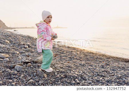 Cute child in winter clothes playing on a rocky beach at sunset 132947752