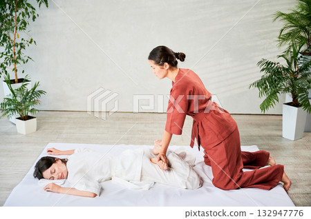 Masseuse making thai yoga massage. Woman therapist doing traditional massage treatment, stretching female patient's body. Client lying on mat, enjoying hand massage in calm atmosphere. 132947776