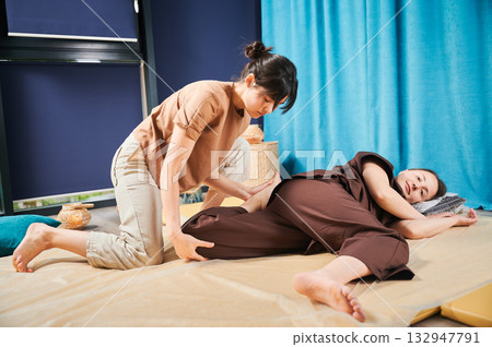 Masseuse making thai yoga massage. Woman therapist doing traditional massage treatment, stretching female patient's body. Client lying on mat, enjoying hand massage in calm atmosphere. 132947791