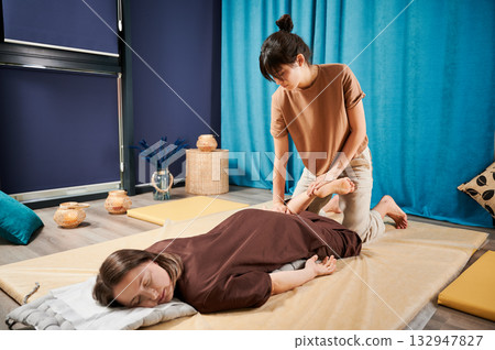 Masseuse making thai yoga massage. Woman therapist doing traditional massage treatment, stretching female patient's body. Client lying on mat, enjoying hand massage in calm atmosphere. 132947827