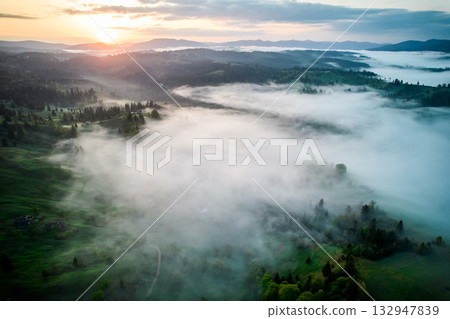 Aerial view of sunrise over mist-covered landscape, with golden light illuminating rolling hills and valleys. Thick fog creates mystical atmosphere, partially obscuring lush greenery and trees below. 132947839