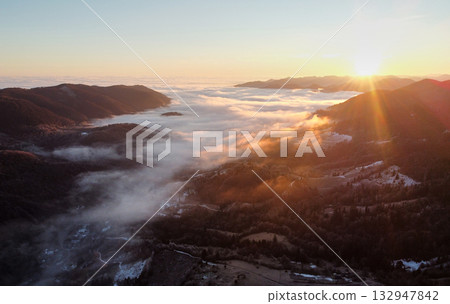 Setting sun casts golden hue over vast sea of clouds, blanketing valleys between forested hills and distant mountain ranges. Aerial view of tranquil scene captures serene beauty of nature. Setting sun casts golden hue over vast sea of clouds, blanketing valleys between forested hills and distant mountain ranges. Aerial view of tranquil scene captures serene beauty of nature. 132947842