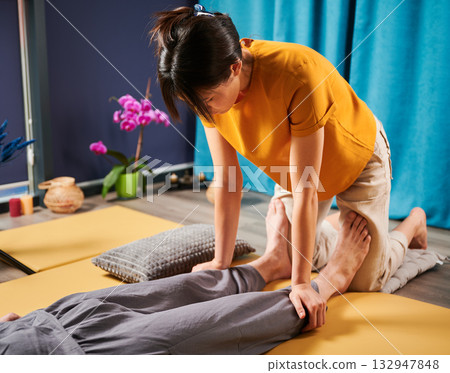 Masseuse making thai yoga massage. Woman therapist doing traditional massage treatment for male patient indoors. Client lying on mat, meditating, enjoying hand massage in calm atmosphere. Masseuse making thai yoga massage. Woman therapist doing traditional massage treatment for male patient indoors. Client lying on mat, meditating, enjoying hand massage in calm atmosphere. 132947848