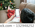 Boy in red plaid pajama bottoms and a t-shirt is lying on sofa, holding a smartphone with blank white screen with decorated Christmas tree in background. Holiday social media or technology themes 132948252