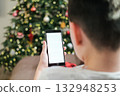 Rear view close-up of boy's hands holding smartphone with blank white screen, focused against blurred background of Christmas tree with glowing lights. Holiday-themed app or social media mockup 132948253