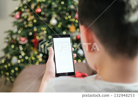 Rear view close-up of boy's hands holding smartphone with blank white screen, focused against blurred background of Christmas tree with glowing lights. Holiday-themed app or social media mockup Rear view close-up of boy's hands holding smartphone with blank white screen, focused against blurred background of Christmas tree with glowing lights. Holiday-themed app or social media mockup 132948253