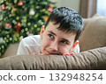 Portrait of cute nine-year-old boy looking up dreamily and smiling slightly against backdrop of Christmas tree and glowing lights in anticipation of holiday and gifts 132948254