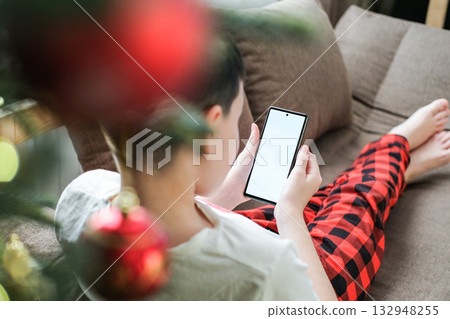 Over-the-shoulder shot of boy in red pajamas lying on sofa using smartphone with blank white screen with blurred Christmas ornament in foreground. Perfect for holiday-themed digital mockups Over-the-shoulder shot of boy in red pajamas lying on sofa using smartphone with blank white screen with blurred Christmas ornament in foreground. Perfect for holiday-themed digital mockups 132948255