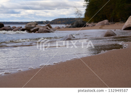 Low angle view of small waves reaching sandy beach with rocks and trees. 132949587