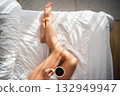 Close-up of female legs with cup of coffee on bed. 132949947