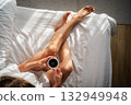 Close-up of female legs with cup of coffee on bed. 132949948