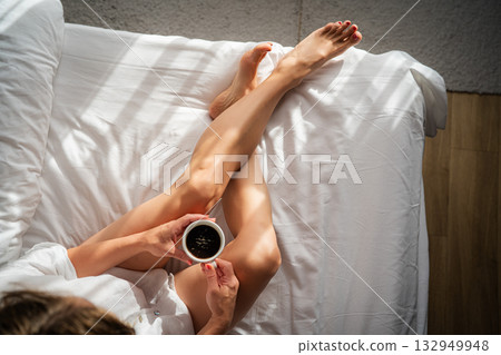 Close-up of female legs with cup of coffee on bed. Close-up of female legs with cup of coffee on bed. 132949948
