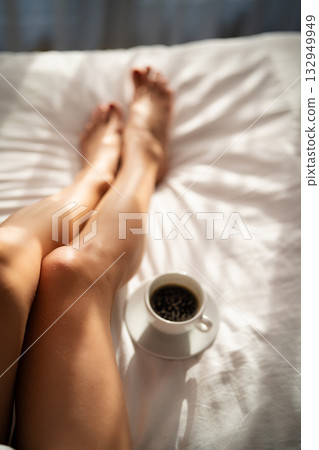 Close-up of female legs with cup of coffee on bed. 132949949