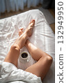 Close-up of female legs with cup of coffee on bed. 132949950