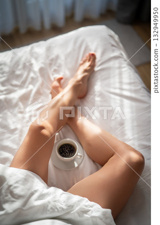 Close-up of female legs with cup of coffee on bed. Close-up of female legs with cup of coffee on bed. 132949950