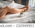 Close-up of female legs with cup of coffee on bed. 132949951