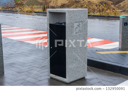 A sleek, gray concrete trash bin stands on a wet urban street, with a nearby pedestrian crossing and overcast sky 132950091