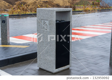 A stylish waste bin stands on a wet city street, showcasing a blend of stone and metal design amidst a clean urban environment 132950092