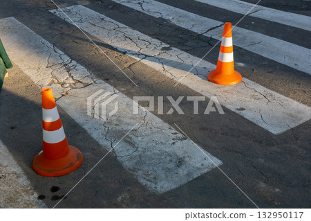 Two orange traffic cones are placed next to a pedestrian crosswalk on a cracked pavement as shadows stretch in the afternoon light Two orange traffic cones are placed next to a pedestrian crosswalk on a cracked pavement as shadows stretch in the afternoon light 132950117