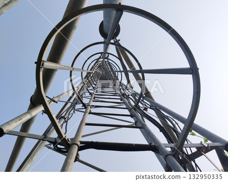 Upward view of steel tower with circular safety rings and climbing ladder, industrial iron structure against clear blue sky, engineering design for telecommunications or high-rise installation 132950335