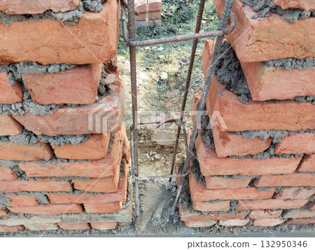 Unfinished concrete pillar structure with exposed steel rods between two brick walls in rural construction site, showing early stage of column reinforcement without concrete filling 132950346