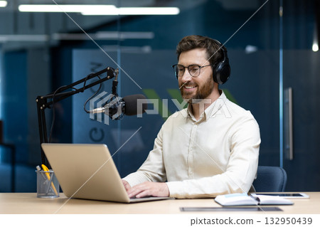 Smiling young man in headphones sitting at a desk in the office in front of a screen with a microphone and working on a laptop. Smiling young man in headphones sitting at a desk in the office in front of a screen with a microphone and working on a laptop. 132950439