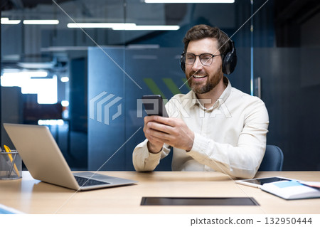 Smiling young man working in the office, sitting at his desk wearing headphones and using his mobile phone. Smiling young man working in the office, sitting at his desk wearing headphones and using his mobile phone. 132950444