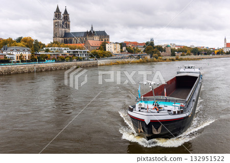 Cargo barge ship sails river Elbe historic Magdeburg city skyline cathedral Dom towers and colorful autumn trees. Overcast sky calm water create tranquil atmosphere European river inland transport Cargo barge ship sails river Elbe historic Magdeburg city skyline cathedral Dom towers and colorful autumn trees. Overcast sky calm water create tranquil atmosphere European river inland transport 132951522