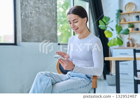 Young smiling woman sitting at home on chair using smartphone 132952407