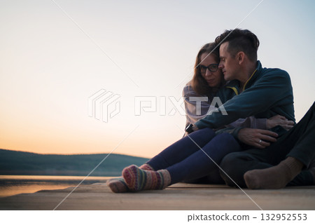 Couple Sitting by Lake at Sunset Embracing Couple Sitting by Lake at Sunset Embracing 132952553