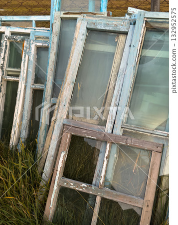 Weathered window frames lean in overgrown grass with peeling blue paint and sunlight Weathered window frames lean in overgrown grass with peeling blue paint and sunlight 132952575