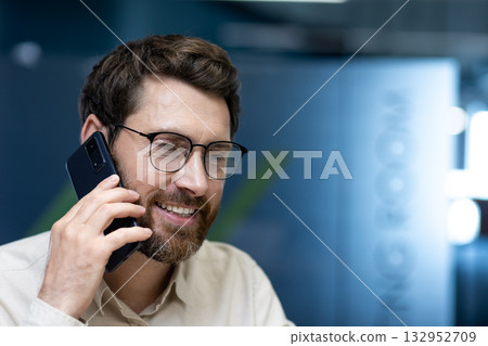 Close-up photo of a young man in glasses and with a beard talking on the phone at his workplace in the office. 132952709