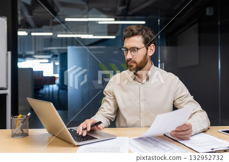 A concentrated young man is sitting in the office at a desk, holding documents in his hands and working on a laptop. A concentrated young man is sitting in the office at a desk, holding documents in his hands and working on a laptop. 132952732