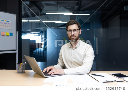 Portrait of a young man working in an office, sitting at a desk with a laptop and looking intently at the camera. Portrait of a young man working in an office, sitting at a desk with a laptop and looking intently at the camera. 132952748