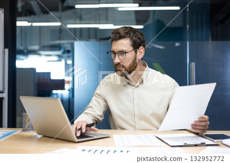 Focused young man financial specialist and analyst working in office with papers and documents, using laptop. Focused young man financial specialist and analyst working in office with papers and documents, using laptop. 132952772