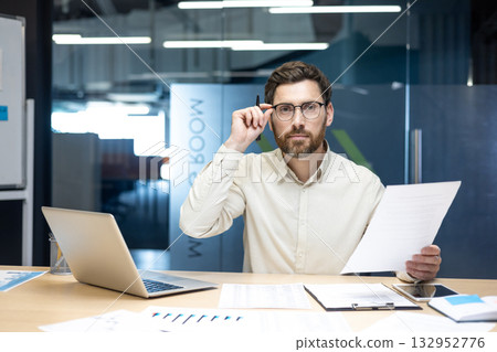 Portrait of a young man working in an office with documents, sitting at a table, supporting his glasses with his hand, looking at the camera. Portrait of a young man working in an office with documents, sitting at a table, supporting his glasses with his hand, looking at the camera. 132952776