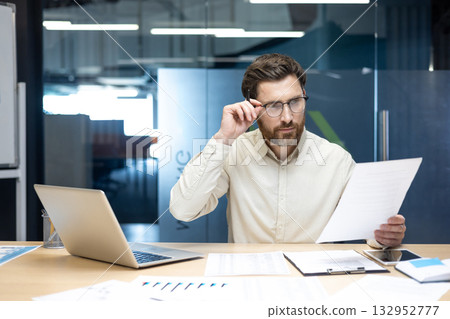 Office worker, young businessman sitting at a desk with a laptop, supporting his glasses with his hand and looking at documents. Office worker, young businessman sitting at a desk with a laptop, supporting his glasses with his hand and looking at documents. 132952777