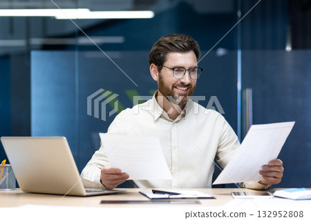 A young businessman sits in the office at a desk with a laptop and looks at the documents he holds in his hands, conducting analysis and examination. 132952808