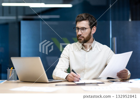 Concentrated male banker and office worker sitting in the office at the table, working on a laptop and with documents. 132952841