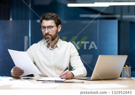 A serious young male businessman is sitting in an office at a desk, working on a laptop and with documents. 132952843
