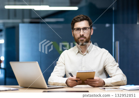 Portrait of a young successful serious male lawyer, businessman sitting at a table with a laptop in the office, holding a notebook and a pen in his hands and looking confidently at the camera. Portrait of a young successful serious male lawyer, businessman sitting at a table with a laptop in the office, holding a notebook and a pen in his hands and looking confidently at the camera. 132952851
