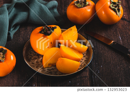 Pieces and halves of persimmon fruit on a plate and on a table Pieces and halves of persimmon fruit on a plate and on a table 132953023