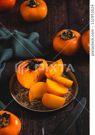 Pieces and halves of persimmon fruit on a plate and on a table vertical view 132953024