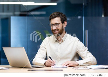A smiling young man sits in the office at a desk in front of a notebook, makes notes in a notebook and looks at the screen with a smile. A smiling young man sits in the office at a desk in front of a notebook, makes notes in a notebook and looks at the screen with a smile. 132953619