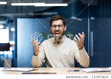 Close-up portrait of an angry and irritated young man sitting in an office at a desk and yelling at the camera while waving his hands. 132953624