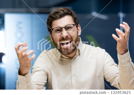 Close-up portrait of a young man in glasses and a shirt who is in the office and angrily talking and shouting at the camera while gesturing with his hands. 132953625