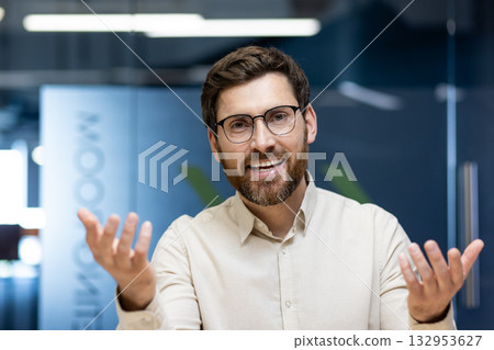 Close-up portrait of a young man sitting in an office at a desk, talking and gesturing to the camera. 132953627