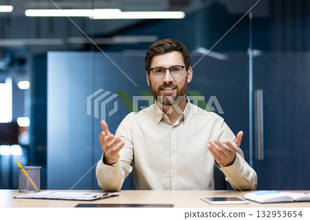 Portrait of a young man sitting in the office at a table in front of the camera and talking on a video call, gesturing to the camera. Portrait of a young man sitting in the office at a table in front of the camera and talking on a video call, gesturing to the camera. 132953654