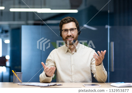 A smiling young man wearing a headset is sitting at a desk in the office and talking to the camera remotely gesturing with his hands. 132953877