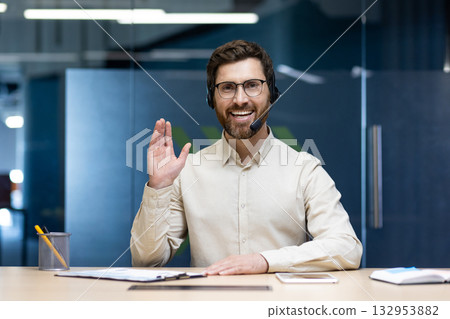 Portrait of a happy and successful young man in a headset sitting at a table in the office in front of the camera, smiling online, greeting and waving. 132953882